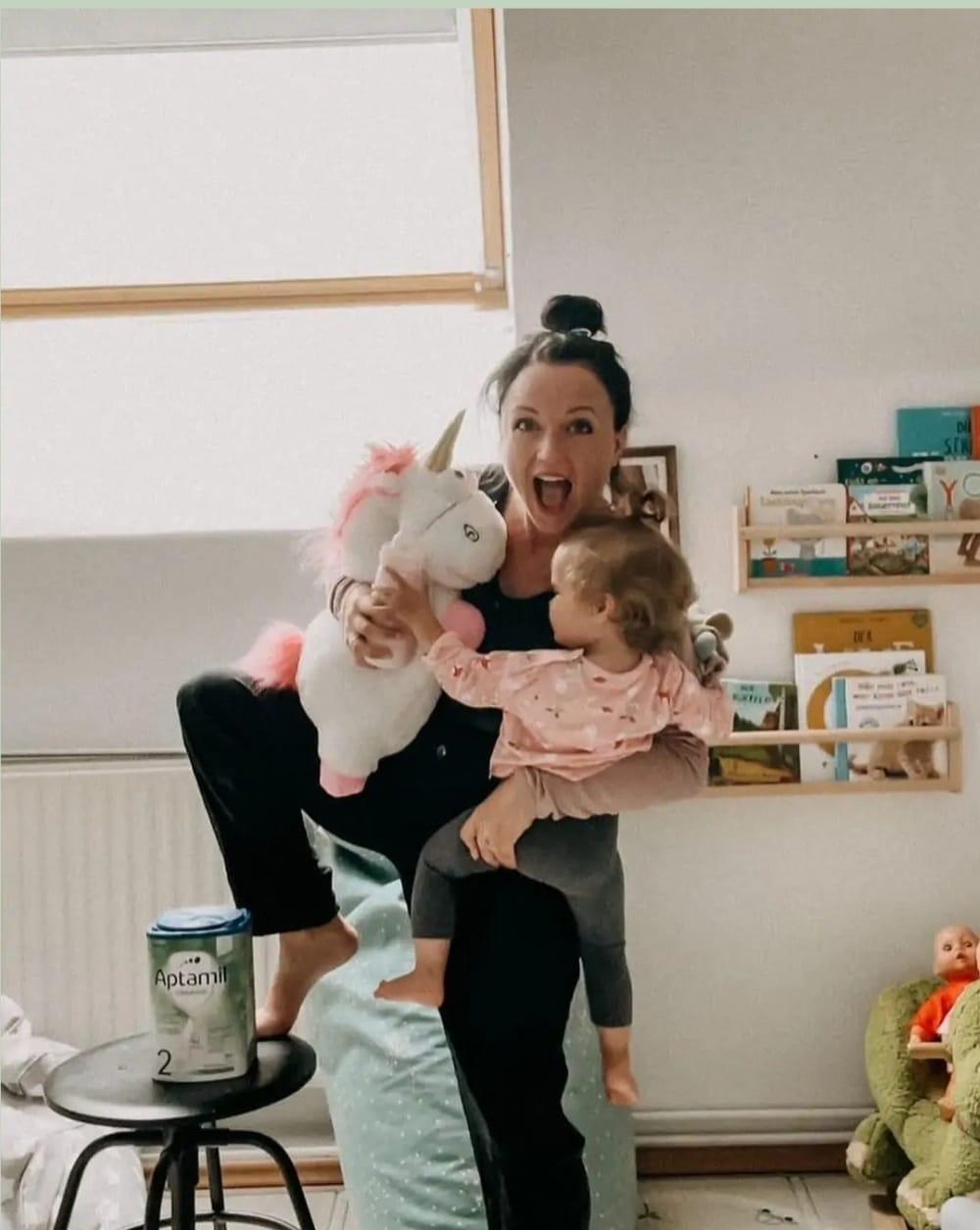Julia mit Tochter und Plüscheinhorn. Sie wirkt ausgelassen.